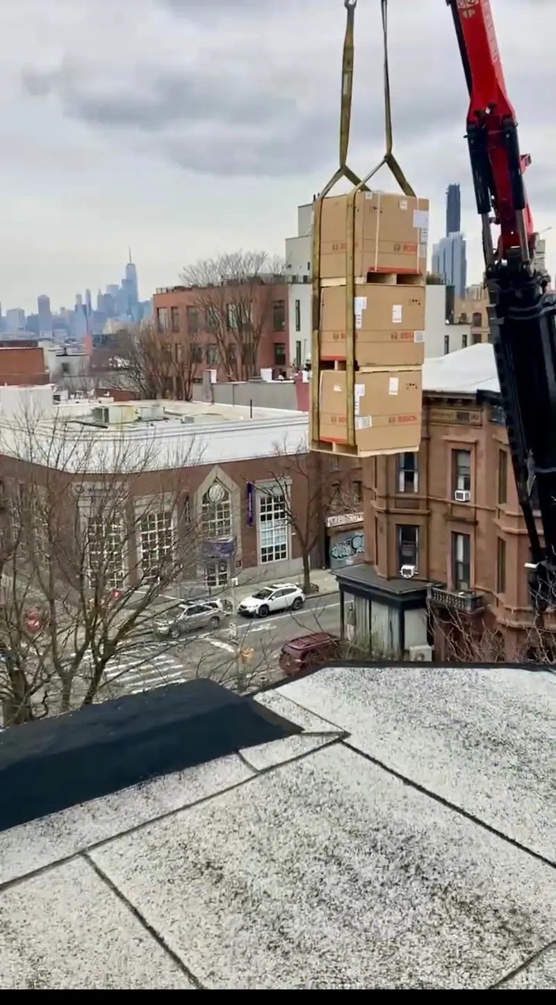 Three boxed Bosch HVAC units lifted by crane onto a Brooklyn brownstone roof with Lower Manhattan in the distance