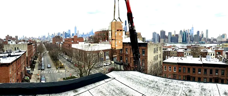 Crane hoisting boxed HVAC units over Park Slope rooftops with the Manhattan skyline and Brooklyn Bridge behind