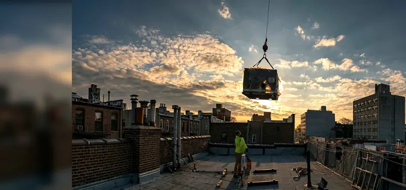 Rooftop HVAC unit suspended from a crane at sunrise while a Brownstone technician in a hi-vis jacket watches