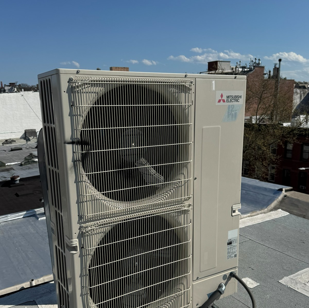 Mitsubishi Electric outdoor condensing unit on a Brooklyn rooftop, showing clear MITSUBISHI ELECTRIC branding and Manhattan skyline in the background